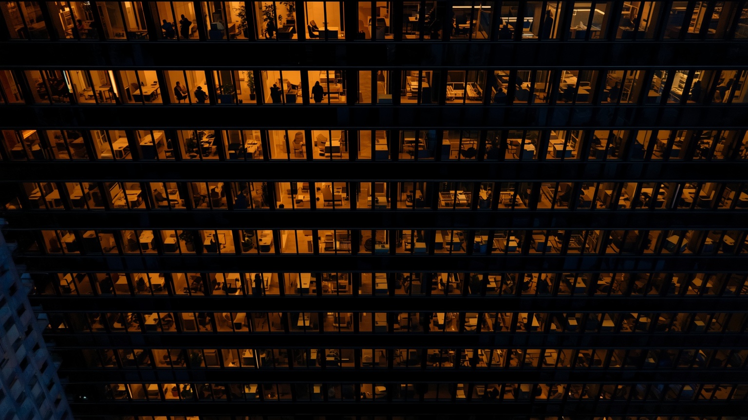 A corporate campus at dusk, with orderly rows of illuminated office windows casting amber light